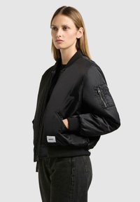 Schwarze Bomberjacke mit Reißverschluss vorne, gerippten Bündchen und seitlichen Taschen. Verfügt über eine Reißverschlusstasche am Ärmel und ein Etikett am Saum.