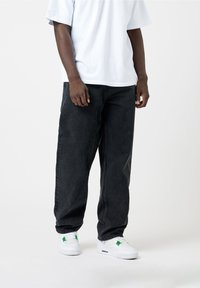 Schwarze Denim-Hose mit lockerem Schnitt, kombiniert mit einem weißen T-Shirt mit Rundhalsausschnitt. Die Schuhe sind weiße Sneaker mit grünen Akzenten.