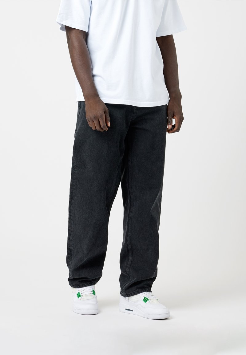 Schwarze Denim-Hose mit lockerem Schnitt, kombiniert mit einem weißen T-Shirt mit Rundhalsausschnitt. Die Schuhe sind weiße Sneaker mit grünen Akzenten.