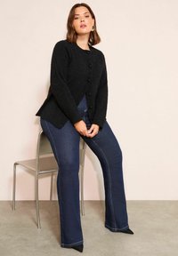 Frau trägt einen schwarzen, geknöpften Cardigan, dunkelblaue ausgestellte Jeans und schwarze spitze Schuhe, sitzt auf einem Metallstuhl vor schlichter Hintergrund.