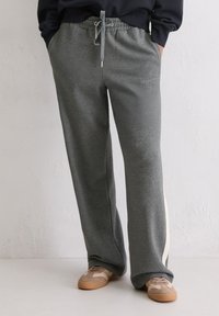 Persoon in grijze joggingbroek met trekkoord en zijstreep, gecombineerd met beige sneakers, staand tegen een lichte gestructureerde muur.