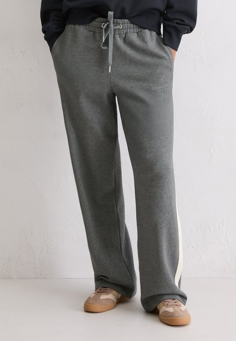 Persoon in grijze joggingbroek met trekkoord en zijstreep, gecombineerd met beige sneakers, staand tegen een lichte gestructureerde muur.