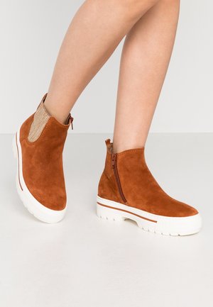 Brune ruskindsankelstøvler med et tekstureret stofpanel, lynlås i siden og hvid gummisål med en kontrasterende orange stribe.