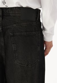 Black denim jeans featuring a back patch, visible stitching, and a single back pocket. The fabric has a textured finish with a slight sheen.