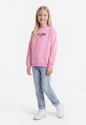 Jeune fille aux longs cheveux blonds souriante, portant un sweat-shirt rose Nike, un jean bleu clair et des baskets blanches, debout sur un fond blanc.