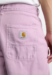 Pink denim pants with a herringbone pattern, featuring a back pocket adorned with a grey Carhartt label and orange logo accent.