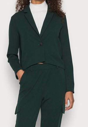 Woman wearing a dark green buttoned blazer over a white turtleneck sweater, paired with matching cargo pants, hands in pockets.