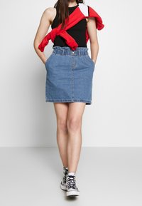 Denimová sukně s nařaseným pasem, zapínáním na knoflík a předními kapsami; doplněná černým tílkem a červeným svetrem přehozeným přes ramena.