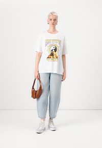 White short-sleeve T-shirt with a colorful graphic featuring a yin-yang symbol, paired with light blue baggy denim jeans and white sneakers.