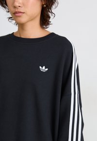 Crna majica s kapuljačom napravljena od mješavine pamuka, sadrži bijeli adidas logo na lijevoj strani prsnog koša i tri bijele pruge duž rukava. Okrugli izrez, opušten fit.