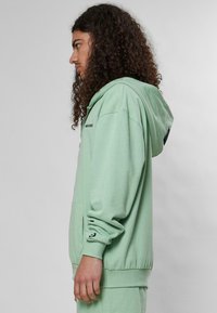 Svijetlozelena hoodie izrađena od mekog materijala, s prednjim džepom, rebrastim manžetama i kapuljačom sa šniranjem. Uključuje logo na prsima.
