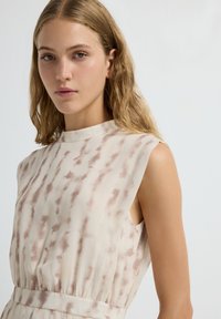 Vestido sin mangas en un ligero color beige con un sutil patrón de tie-dye. Presenta un escote alto y una cintura fruncida para un ajuste relajado. Textura de tela suave.