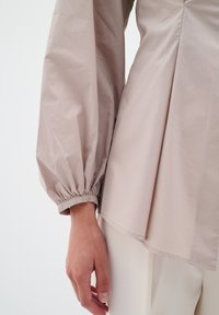 Blusa beige pallido con un corpetto aderente e maniche voluminose. Le maniche hanno polsini elasticizzati e il tessuto appare liscio con una leggera lucentezza.