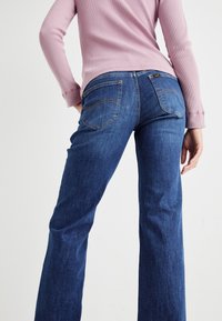 Jeans a palazzo in denim blu scuro con tasche posteriori e dettagli di cucitura in evidenza, abbinati a una maglietta a maniche lunghe di un rosa chiaro.