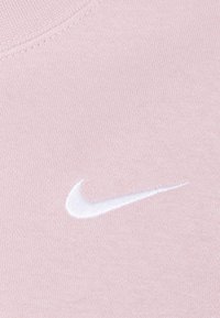 Jasnoróżowa bawełniana tkanina o gładkiej fakturze ma biały haftowany logotyp Nike w dolnym prawym rogu.