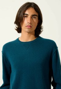 Pull en cachemire teal avec un col rond, des accents côtelés aux épaules, une texture douce et des manches longues. Design lisse et ajusté.