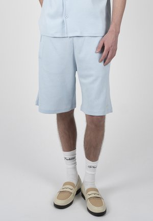 Mann trägt hellblaue, strukturierte Shorts, weiße Socken mit schwarzem Text und beige gewebte Loafers, steht vor einem einfarbigen Hintergrund.