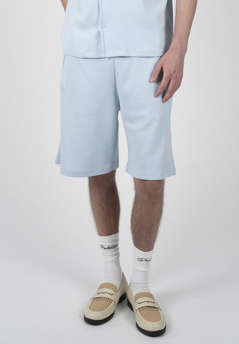 Homme portant un short texturé bleu clair, des chaussettes blanches avec texte noir et des mocassins tissés beige, debout devant un fond uni.