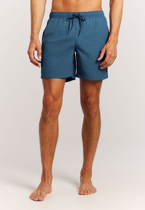 Man met blote benen, dragend blauwe zwembroek met elastische tailleband en zwarte trekkoord, staand op een lichte achtergrond.