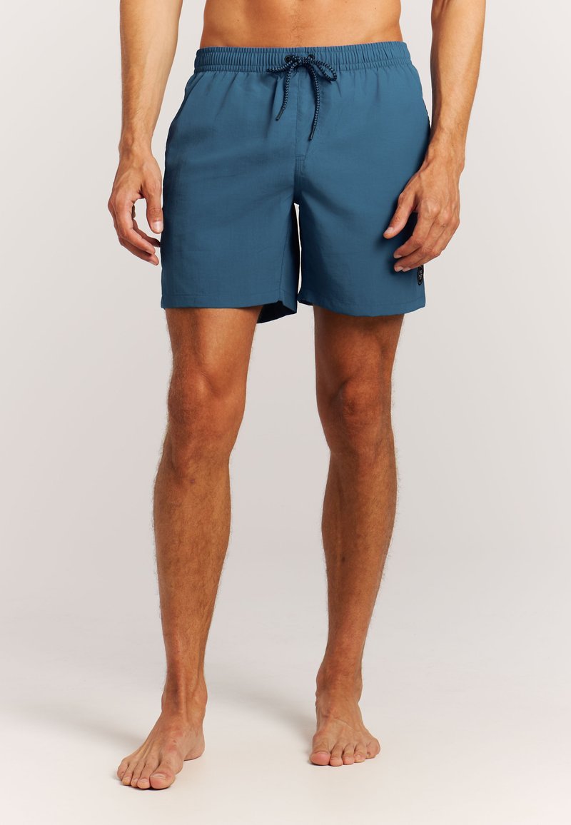 Mand med bare ben iført blå badebukser med elastik i taljen og sort snor, stående på en lys baggrund.