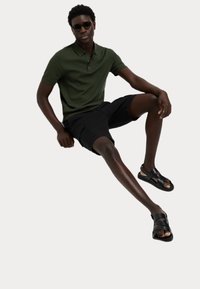 Polo verde, pantaloni neri e sandali neri. Vestibilità slim, tessuto testurizzato, maniche corte e dettaglio con chiusura a due bottoni.