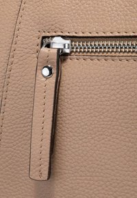 Beige textielachtig leer met een zilveren rits met trekker en stikseldetails. De rits heeft een klein, rechthoekig leren label.