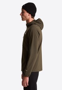 Homme portant une veste à capuche vert olive, un bonnet noir et un pantalon noir, tourné vers la gauche sur un fond blanc uni.