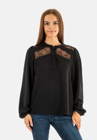 Blusa nera con maniche lunghe, caratterizzata da pannelli in pizzo nella parte superiore del petto e sulle spalle, abbottonatura frontale e polsini arricciati.