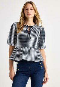 Kvinde iført en marineblå og hvid stribet bluse med pufærmer, peplumkant og to sorte sløjfer ved halsudskæringen, kombineret med mørkeblå jeans.