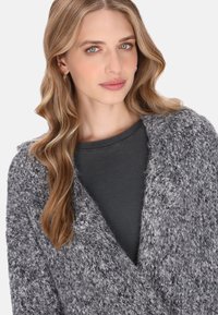 Donna con lunghi capelli biondi ondulati che indossa orecchini a cerchio in argento, una maglietta grigio scuro a coste e un cardigan grigio a maglia con trama.