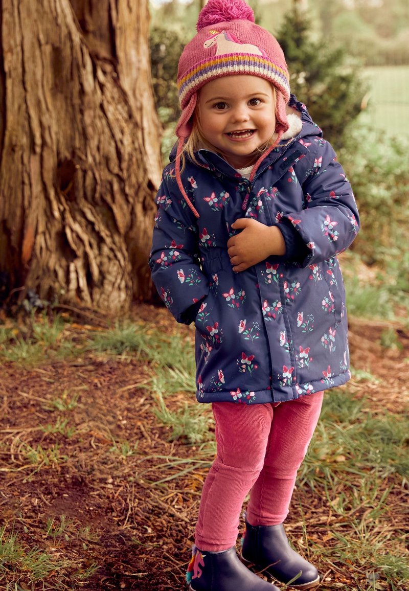 Jachetă puffer florală de marină, colanți roz din cordura și cizme negre cu design de unicorni. Copilul poartă o pălărie roz cu pom-pom și clapete pentru urechi.