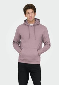 Jonge man met krullend haar, gekleed in een mauve hoodie en zwarte broek, met handen in de voorkantzak, vooruit kijkend tegen een effen lichte achtergrond.