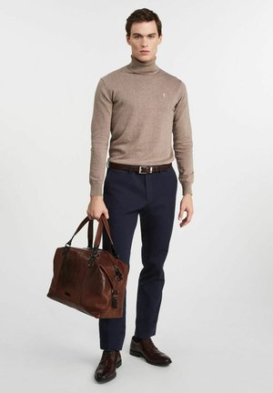 Mężczyzna w beżowym golfie, granatowych spodniach i brązowych butach, trzymający dużą brązową skórzaną torbę podróżną z dwoma uchwytami.