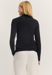 Schwarzes Langarmoberteil mit einem Cowl-Neck, taillierte Passform, glatte Textur und minimalistischen Stil. Kombiniert mit hellen Hosen.