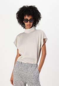 Un haut beige clair à col montant avec des manches courtes, associé à un pantalon gris texturé. Le haut est lisse, créant un look minimaliste.