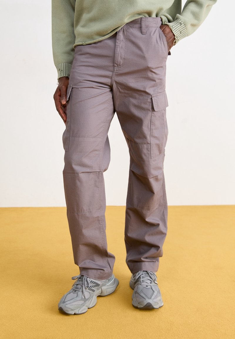 Personne portant un pantalon cargo violet clair et des baskets grises, debout avec une main dans la poche sur un sol jaune.