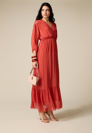 Donna che indossa un lungo vestito rosso con scollo a V e maniche a tre quarti, tiene in mano una borsa beige e indossa sandali metallici con tacco e braccialetti.