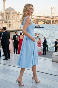 Mujer con vestido sin mangas azul claro y tacones plateados de pie junto a un paseo marítimo con un puente, un edificio histórico y personas con ropa formal.
