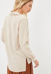 Blusa beige de manga larga con un sutil brillo, pliegue en la parte trasera y aberturas laterales, combinada con una falda marrón estampada. Textura suave.