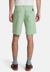 Jasnozielone bawełniane szorty z dwiema tylnymi kieszeniami, ozdobione guzikami i markową naszywką, w zestawieniu z szarymi skarpetkami i brązowymi butami.