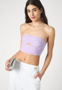 Lilac strapless crop top with a front cutout, smooth texture, and contoured design, paired with high-waisted white trousers and a silver clutch.