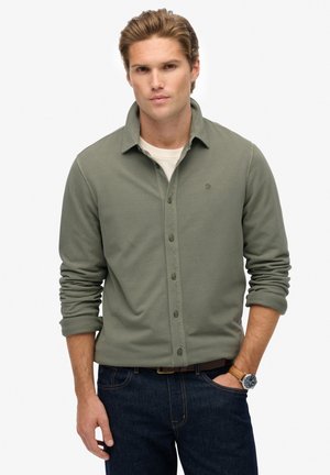 Olijfgroen blouse met lange mouwen en knopen, met een relaxte pasvorm, voorzien van een subtiel logo, een klassieke kraag en opgerolde mouwen. Gepaard met donkere jeans.