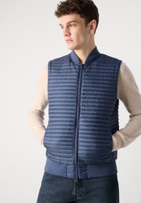 Jūras spēku zila, quiltēta veste ar rāvējslēdzēju priekšā, ribotu apkakli un apakšu. Izgatavota no viegla materiāla ar gludu tekstūru un minimālistisku dizainu.