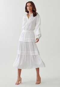 Vestido blanco con cuello abotonado, mangas largas y falda de volantes. Presenta plisados horizontales en la cintura y en la parte inferior de la falda, con una textura de tela suave.