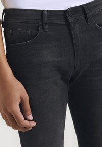 Närbild av en person som bär svarta tajta jeans med synliga framfickor, bältesöglor och en hand avslappnat vid sidan.