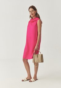 Mouwloze jurk in felroze stof, losse pasvorm, met een kraag. Gecombineerd met een beige handtas en crèmekleurige sandalen met gekruiste banden.