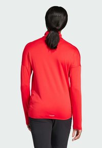 Top rosso a maniche lunghe con collo alto, texture morbida e vestibilità rilassata. Presenta spalle scese e spacchi laterali per facilitare i movimenti.