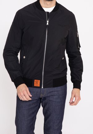 Homme portant une veste bomber noire zippée avec des poignets et un bas côtelés, une étiquette orange à la taille, et un jean bleu foncé sur fond blanc.