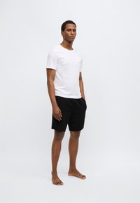 Camiseta blanca de manga corta con un logo rosa en el pecho, combinada con pantalones cortos negros. El modelo está descalzo y posa sobre un fondo neutro.