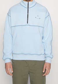 Světle modrá fleecová mikina na zip s vysokým límcem, černým zipem, přední kapsou a kontrastním prošíváním. Malé logo na hrudi.